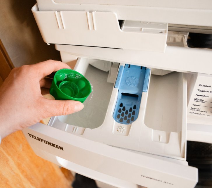 Eine Hand hält eine Verschlusskappe mit Spezialwaschmittel über die geöffnete Einspülkammer einer Waschmaschine.
