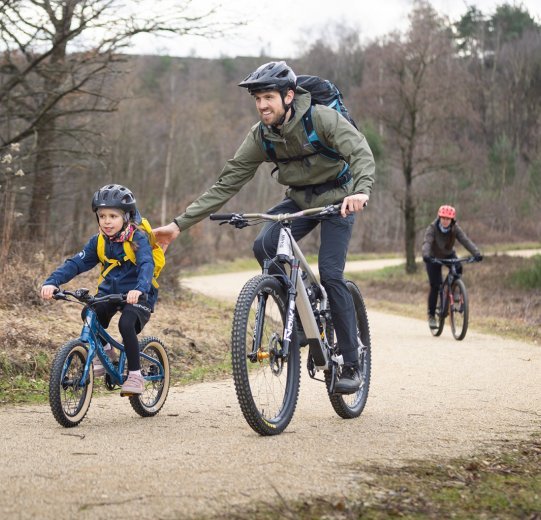 Ein Kind fährt mit einem Erwachsenen Mountainbike. Sie sind auf einem Schotterweg unterwegs.