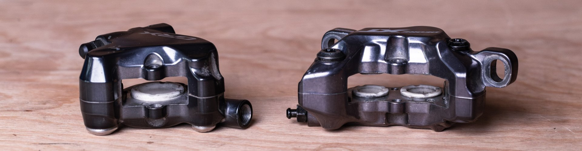 Dos pinzas de freno para frenos de disco de bicicleta están colocadas una al lado de la otra sobre una tabla de madera de tono cálido.