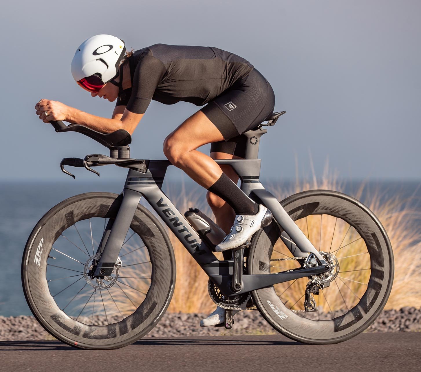 Atleta en posición aerodinámica sobre una bicicleta de contrarreloj con acoples Profile Design, casco aero blanco y ropa de triatlón negra en una carretera costera abierta.