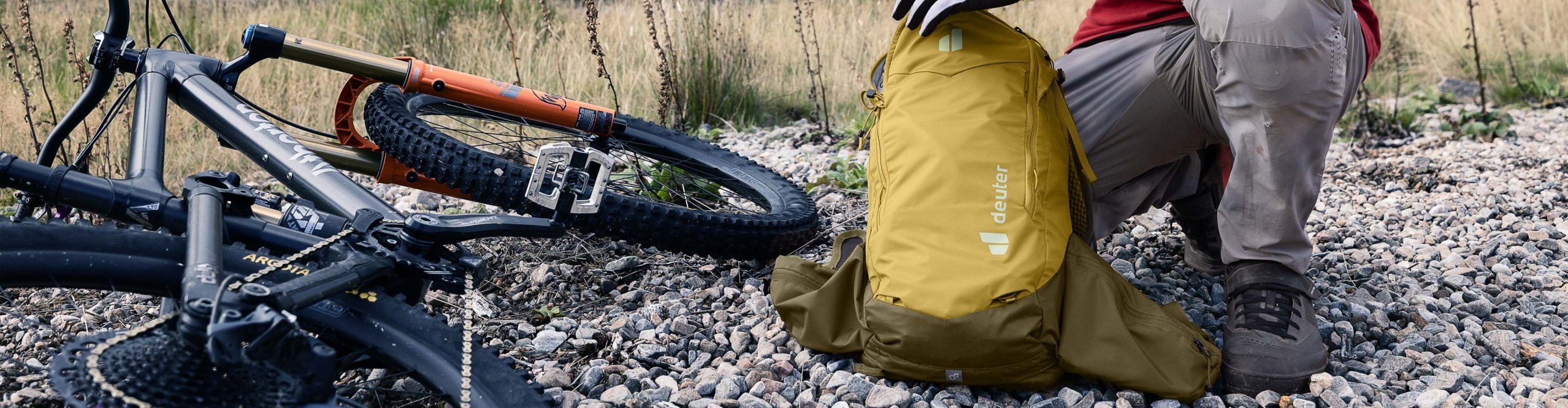 Mountainbiker kniet auf Schotter neben seinem MTB und öffnet einen gelben Deuter Rucksack in alpiner Landschaft