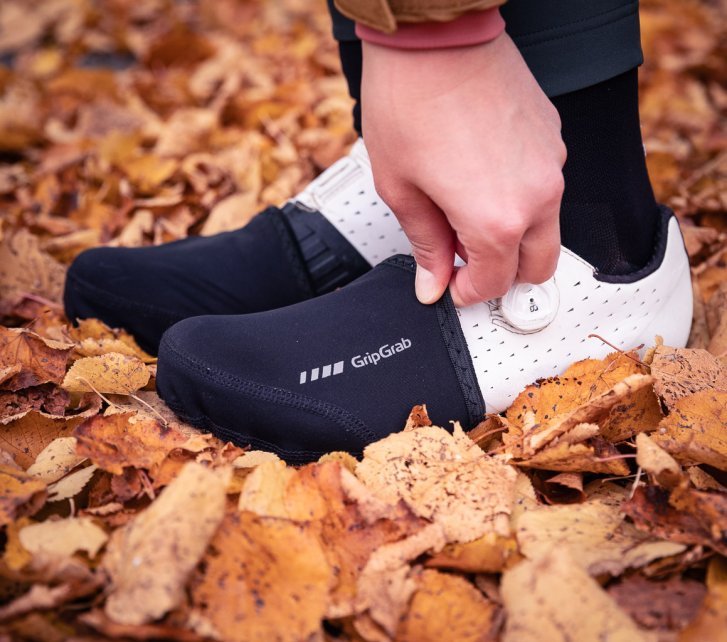 Dos pies con zapatillas gravel blancas y toe caps negras; una mano ajusta la cubierta.