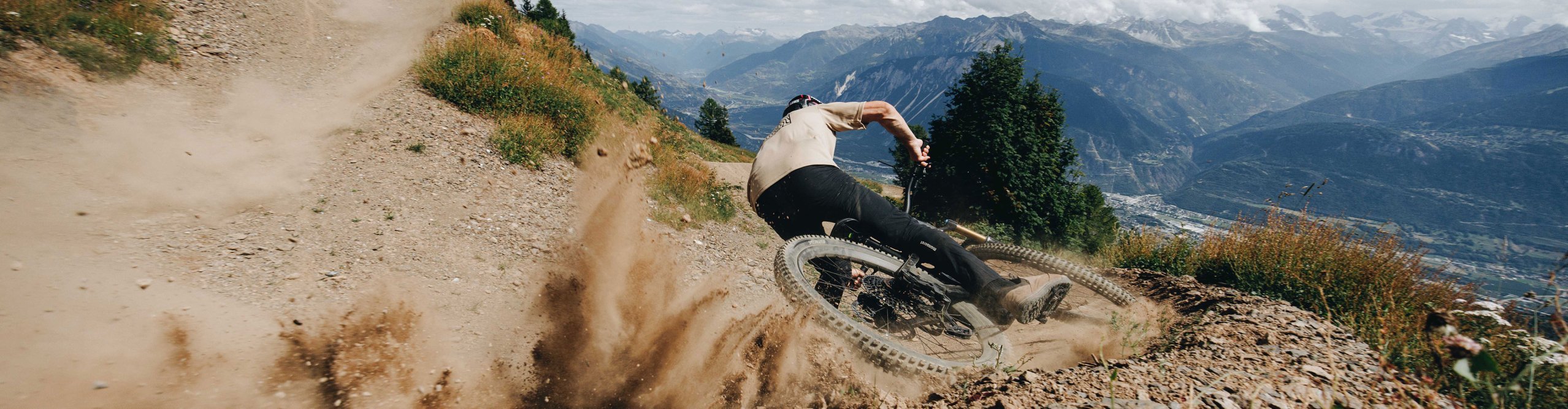 Mountainbiker driftet mit viel Speed durch eine staubige Kurve auf einem alpinen Trail, spektakuläre Bergkulisse im Hintergrund