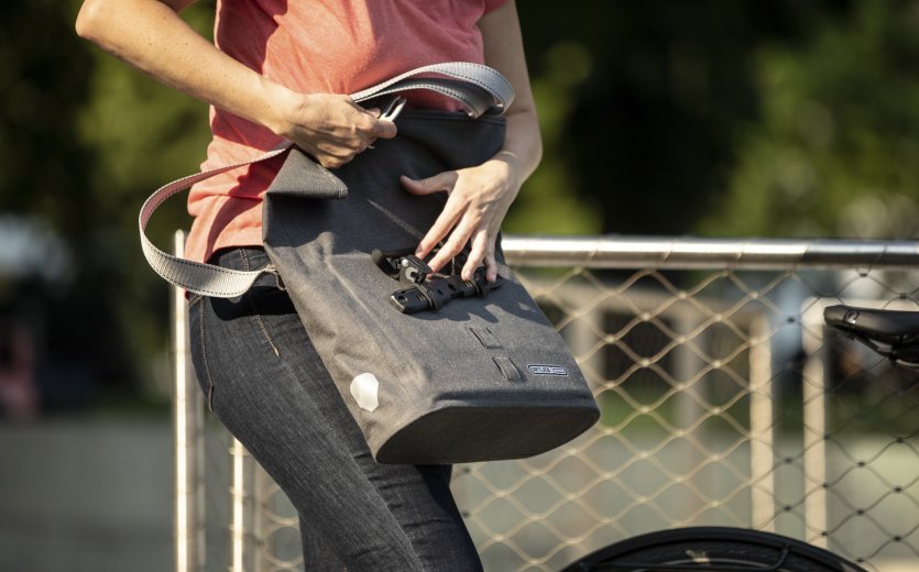 Una persona con una camiseta rosa sujeta una bolsa de bicicleta gris de Ortlieb con una correa para el hombro a una bicicleta. Una valla metálica y un entorno de parque borroso son visibles al fondo.