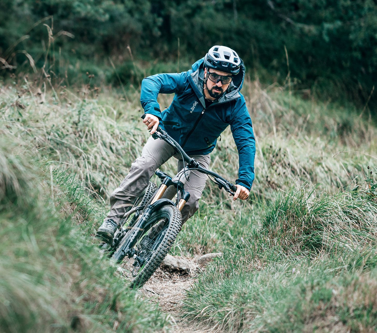 Mountain biker wearing a blue Dirtlej jacket and grey MTB trousers on a narrow trail through a wooded hillside.