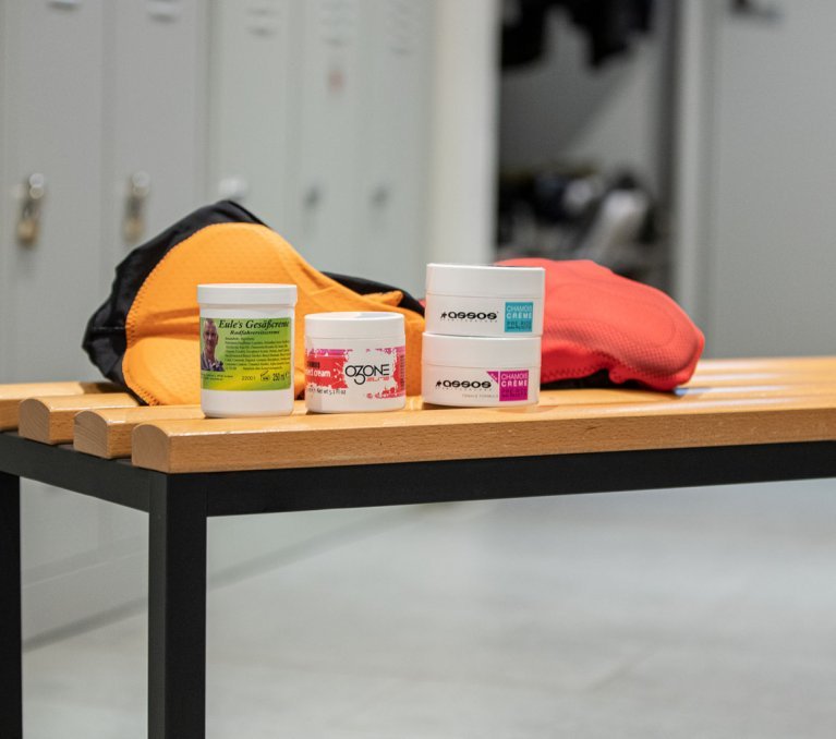 Pictured are 4 varieties of chamois cream from Elite, Assos and Eule in front of 2 bib shorts on a bench in the bc men's locker room.