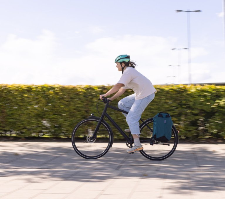 A person rides a bicycle with a helmet along a green hedge, with a large bag on the luggage rack.