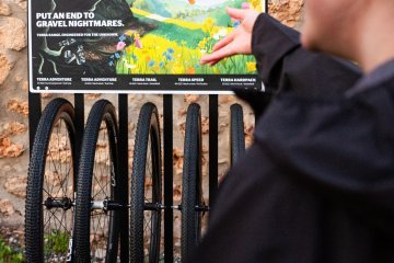 Auswahl verschiedener Continental Gravelreifen in unterschiedlichen Breiten – von Adventure bis Hardpack – präsentiert vor Werbeposter, das die Vielseitigkeit und Performance für jedes Terrain hervorhebt