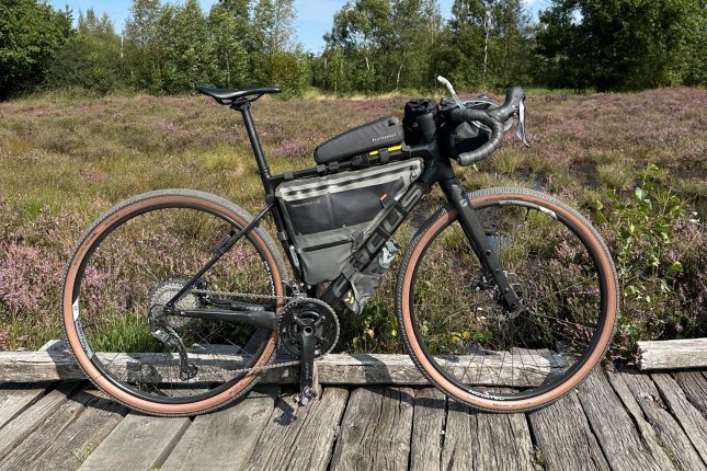 Ein grau-schwarzes Gravel-Bike in Seitenansicht von rechts. Am Fahrrad sind Taschen und ein Trinksystem angebaut. Das Rad steht auf grauen Holzbohlen, dahinter eine blühende Heidelandschaft