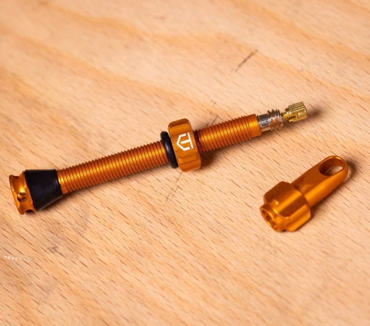 An orange tubeless bicycle valve and an orange valve cap lie on a light wooden surface