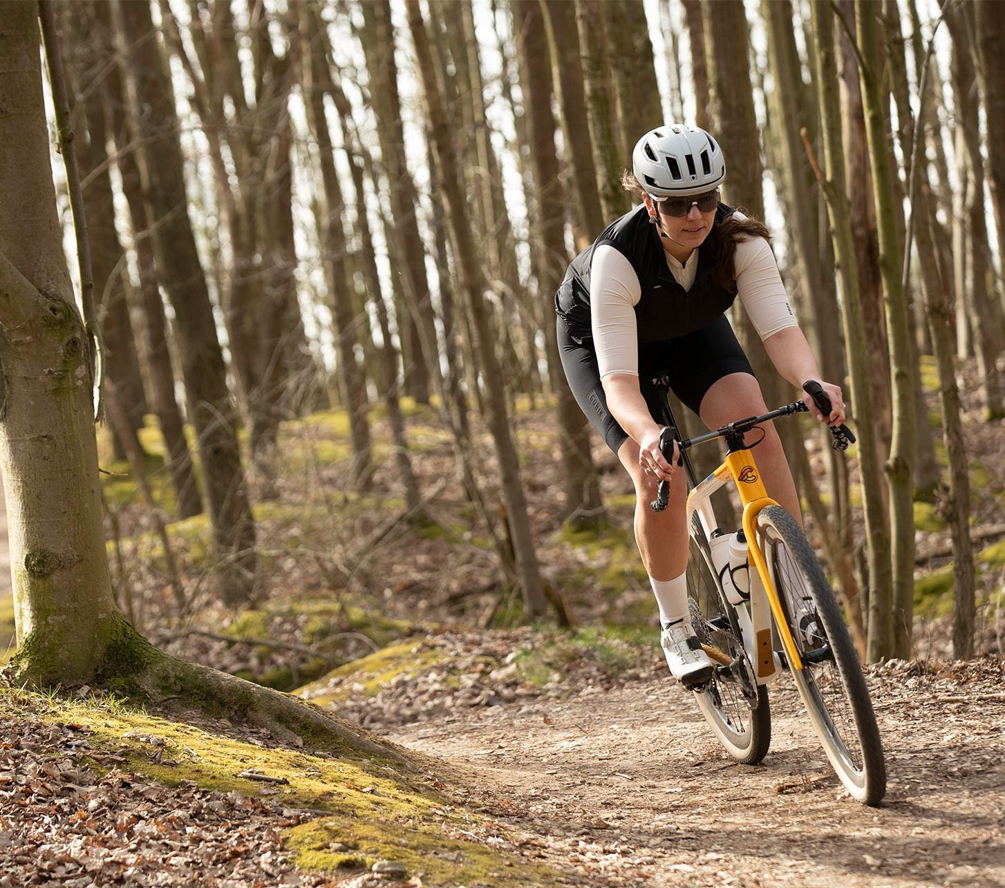 Gravelbikerin fährt mit einem Gravelbike auf einem schmalen Waldtrail durch einen lichten Laubwald. Sie trägt Helm, Sonnenbrille und leichte Fahrradbekleidung für sportliche Fahrten auf Schotter und Naturwegen.