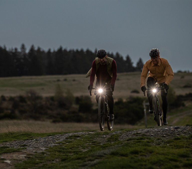 Dos personas montan bicicletas a través de un campo al atardecer. Sus bicicletas tienen luces. El cielo está nublado, el horizonte naranja, con árboles al fondo.