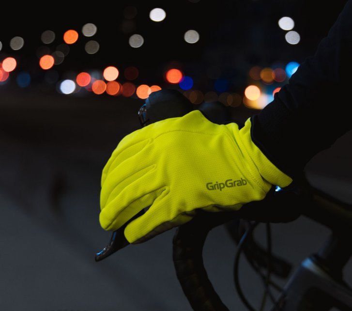 Detailfoto einer Hand in einem neongelben Handschuh der Marke GripGrab an einem Fahrradlenker. Im Hintergrund der nächtlichen Szene sind unscharf bunte Lichter erkennbar.