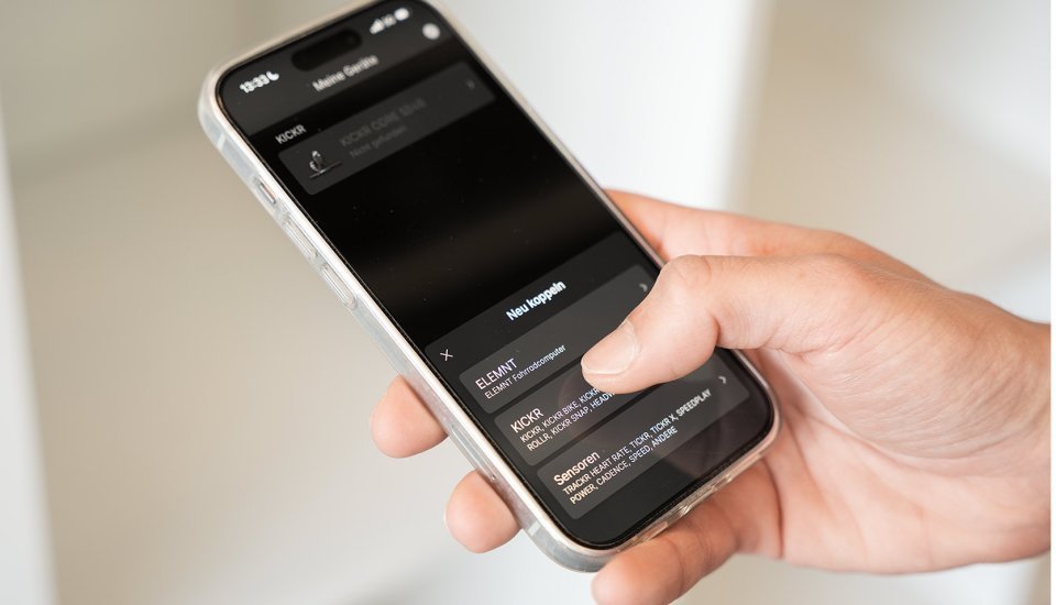 Hand hält ein Smartphone mit geöffneter Wahoo App, in der Wahoo Geräte wie KICKR Rollentrainer und ELEMNT Fahrradcomputer zum Koppeln angezeigt werden.