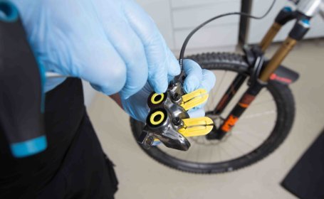 Zwei Hände in blauen Einweghandschuhen setzen die Entlüftungsschraube in einen Bremssattel.
