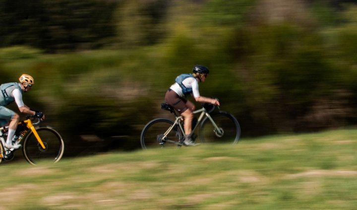 Herbert und Katrin flitzen auf ihren Gravel Bikes durch eine grüne Naturlandschaft. Mitzieheraufnahme von der Seite, nur die beiden Radfahrenden sind scharf abgebildet, der Hintergrund verwischt.