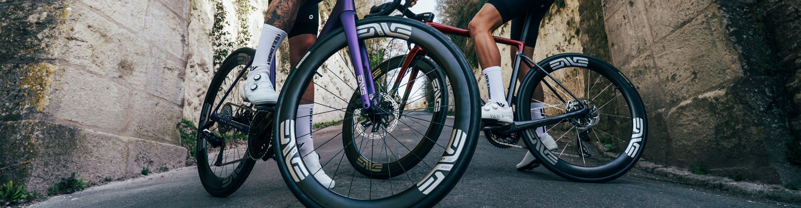 Dos ciclistas de carretera con bicicletas ENVE de alto rendimiento y ruedas aerodinámicas bajo un arco histórico de piedra en una carretera tranquila. Perspectiva dinámica centrada en componentes de carbono premium.