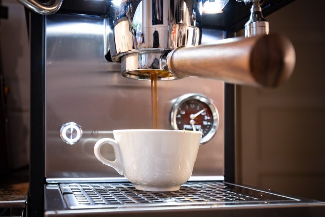 Eine klassische, cremeweiße Cappucchinotasse steht unter einem bodenlosen Siebträger, aus dem nussbraune Crema tropft.