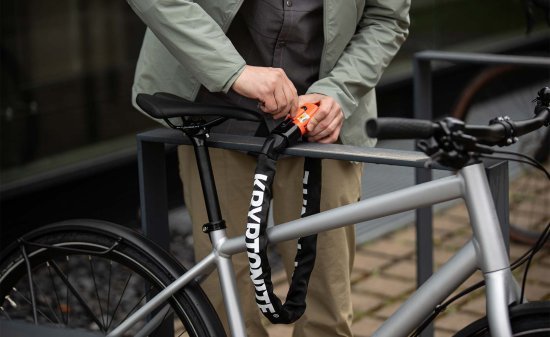 Persona asegurando una bicicleta con un candado de cadena al cuadro y a una barandilla metálica fija para evitar robos.