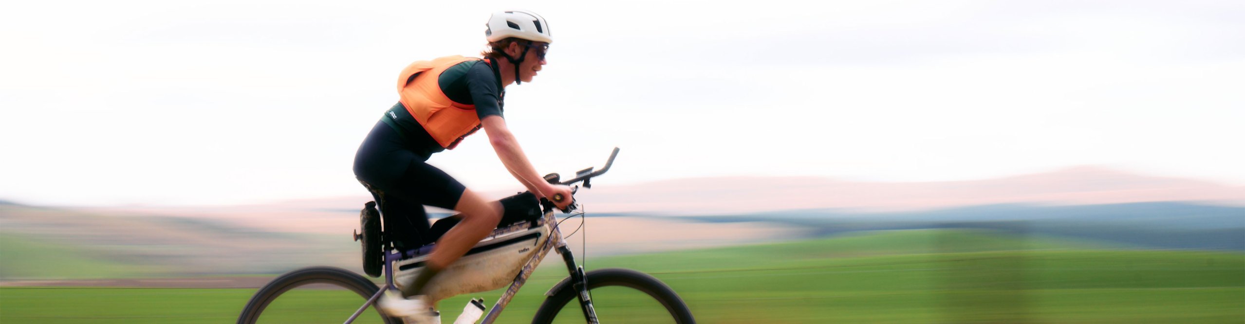 Ciclista deportivo con casco y ropa gravel POC naranja pedaleando rápido en una bicicleta gravel por un camino de tierra, fondo desenfocado que transmite velocidad.