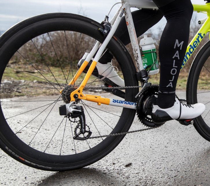 A road cyclist has optimised his bike with CeramicSpeed derailleur pulleys. He rides on a wet street.