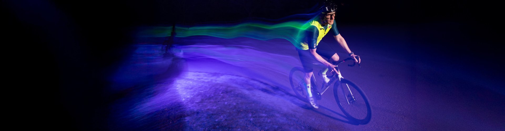 Ein Rennradfahrer mit grellgelber Weste fährt bei Nacht durch einen lilafarbenen Lichtkegel. Er zieht aufgrund der langen Verschlusszeit eine Spur bunter Lichter hinter sich her. 