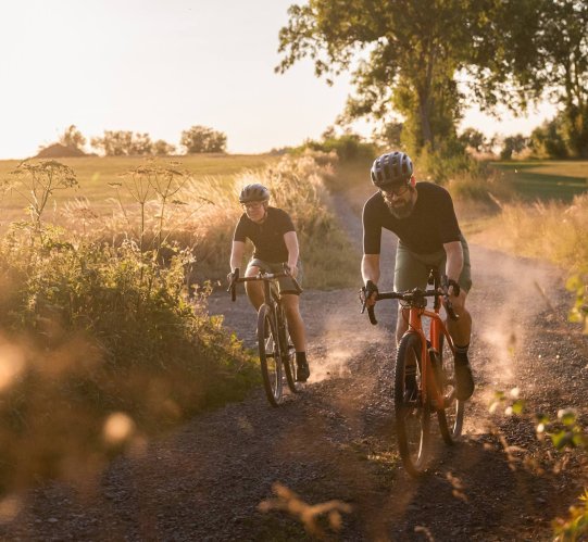 Deux cyclistes gravel roulent sur un chemin poussiéreux au coucher du soleil, poussière visible en contre-jour.