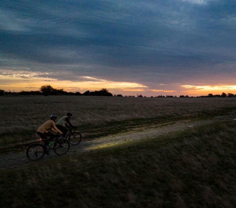 Dos ciclistas con las luces encendidas recorren un camino rural al atardecer.