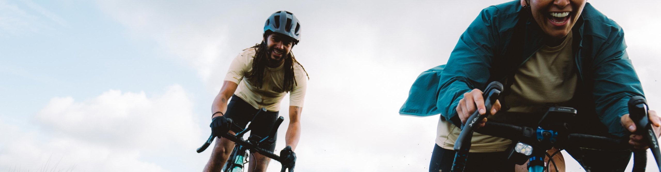 Deux cyclistes gravel avec casque roulent rapidement sur un chemin de gravier mouillé en éclaboussant de l’eau, à vélo gravel avec des vêtements Endura dans un paysage ouvert.