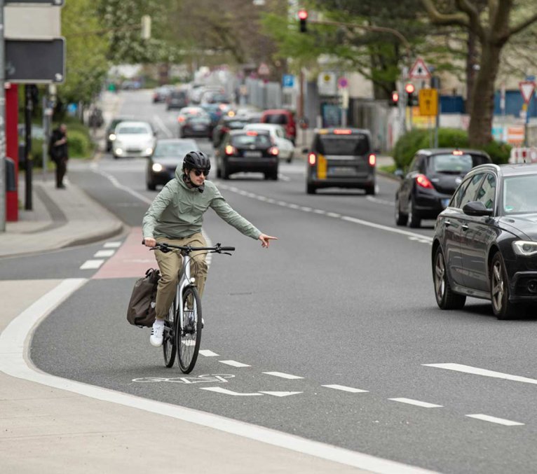 Un cycliste portant un casque roule sur une piste cyclable balisée dans la circulation urbaine et signale d'un geste de la main qu'il change de voie, alors que des voitures circulent sur la route.