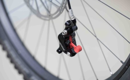 Ein lose an der Bremsleitung hängender SRAM Bremssattel mit eingestecktem Bleed Block. Im Hintergrund unscharf ein MTB-Vorderrad.