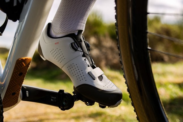 Detailfoto: Ein Fuß mit einem hellgrauen Fahrradschuh auf einem Pedal, unscharf im Vordergrund sehen wir das Unterrohr eines Fahrrads und Teile des Vorderrades.