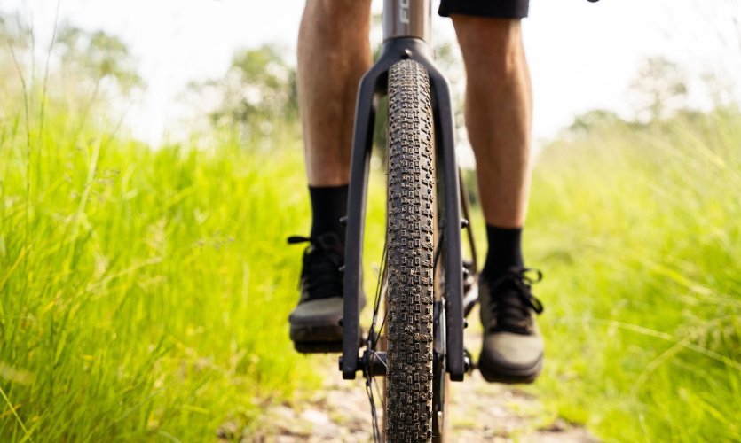 Vue frontale de la roue avant d’un vélo gravel avec pneu à crampons sur un chemin étroit en gravier, entouré d’herbe verte.
