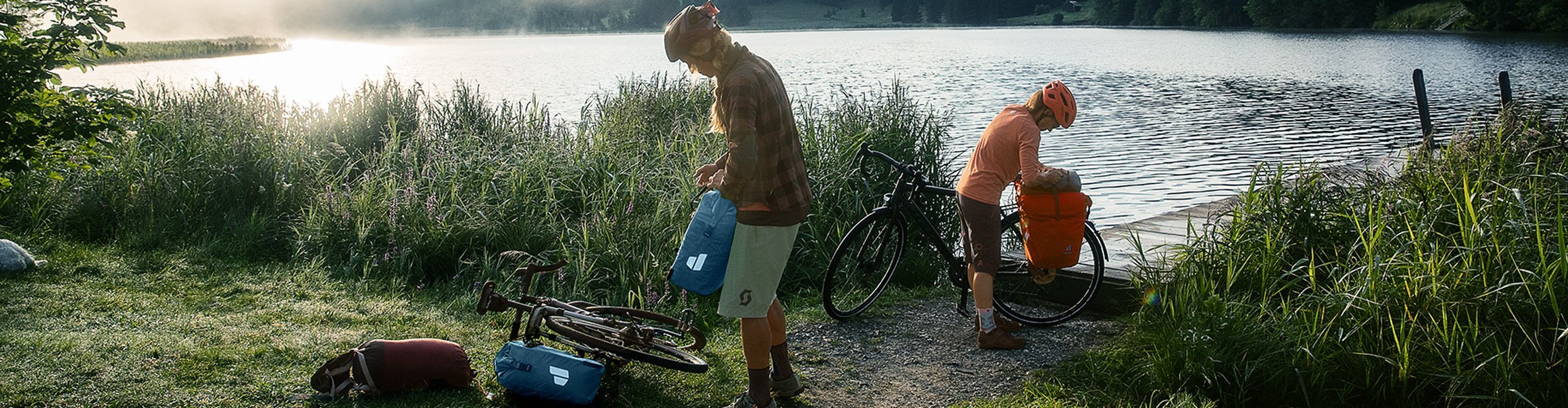 Bikepacking am See bei Sonnenaufgang: Zwei Radfahrer packen wasserdichte Deuter Fahrradtaschen und Gepäckbeutel neben ihren Gravelbikes in alpiner Natur.
