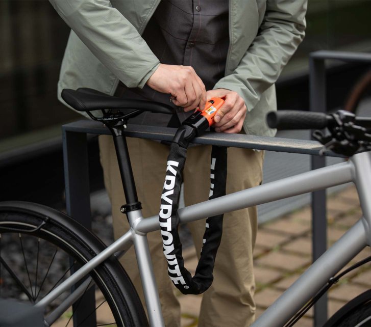 Persona asegurando una bicicleta con un candado de cadena al cuadro y a una barandilla metálica fija para evitar robos.