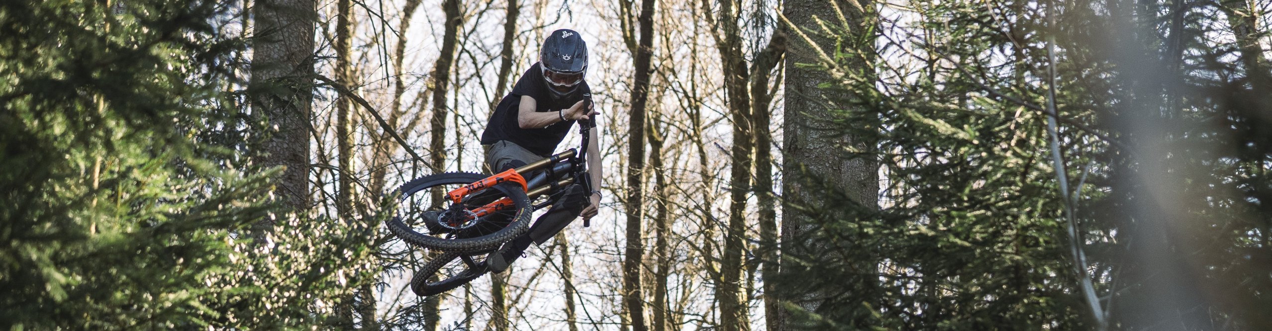 Mountainbiker mit Helm und Schutzkleidung springt über einen großen Holzkicker in einem Wald; das Bike ist in der Luft quer gedreht.