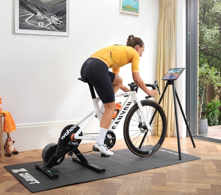 A woman trains on a Wahoo Kickr Core smart trainer with a white Canyon road bike in a bright room with wooden floors.