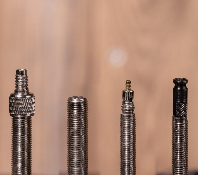 Macro photo: A Dunlop valve, a Schrader valve, a Presta valve and a Schwalbe Clik-Valve side by side in front of a blurred brown wooden surface