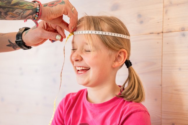 The tattooed hands with colorful nail polish of a mother are holding a measuring tape around the head of a giggling blonde child.