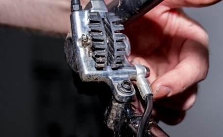 Gros plan sur un étrier de frein de vélo avec ailettes de refroidissement et raccord du câble, tandis qu’une main y travaille.