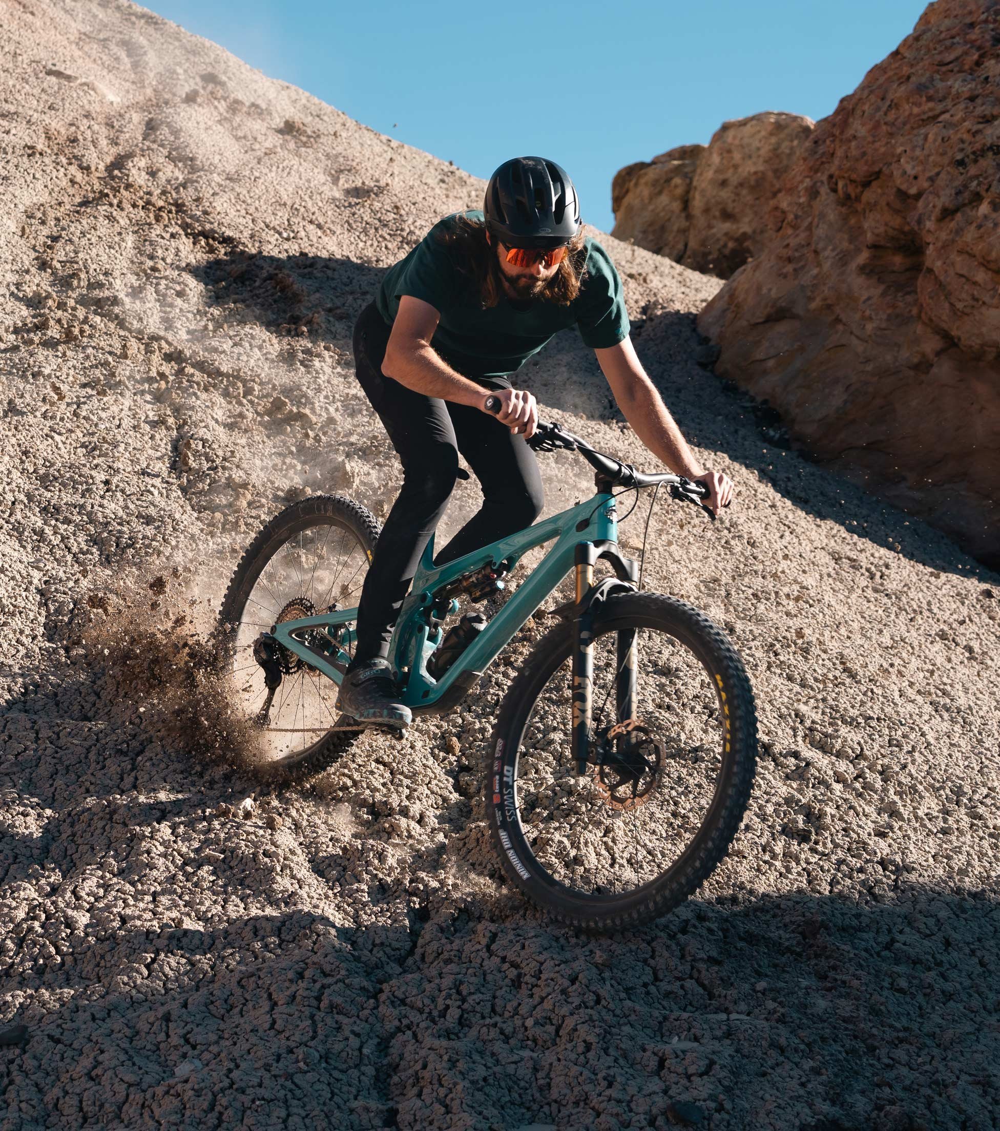 Mountainbiker driftet mit türkisfarbenem Yeti Mountainbike durch steiniges Wüstengelände, Staub spritzt auf.