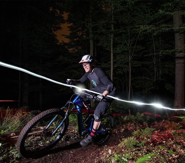 Un ciclista de E-mountain bike pedalea de noche en un sendero del bosque, iluminado por una luz en el casco, con trazos de luz marcando el camino y árboles rodeando el entorno.