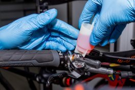Two hands in blue gloves are unscrewing a white funnel with a stopper from the brake lever of a bicycle. The funnel contains a pink fluid used during the bleeding process of a hydraulic disc brake.