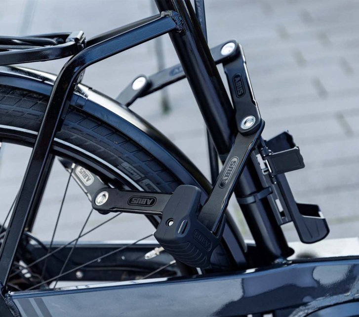 Candado plegable asegurando la rueda trasera y el cuadro de una bicicleta a un punto fijo; fijado de forma compacta al cuadro.