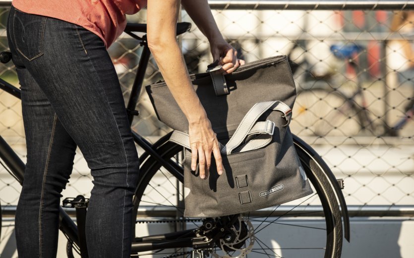 Una persona con una camiseta rosa sujeta una bolsa de bicicleta gris de Ortlieb con una correa para el hombro a una bicicleta. Una valla metálica y un entorno de parque borroso son visibles al fondo.