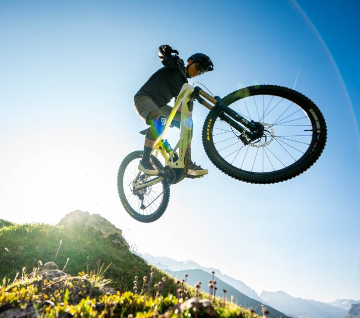A man on an E-MTB jumps a big feature – the bike seems to hover in the air above the camera, blue sky in the background
