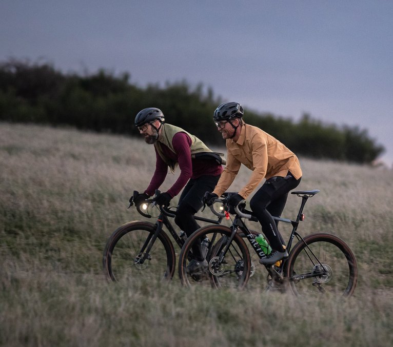 Zwei Radfahrer auf Gravelbikes fahren im Abendlicht durch eine Wiese, ausgestattet mit Helmen und Beleuchtung.