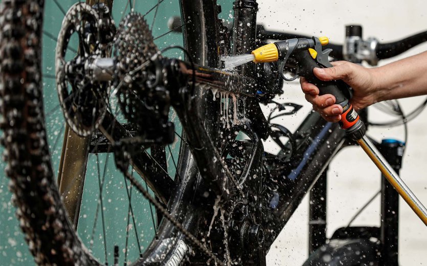 Nahaufnahme eines Fahrradrahmens und Antriebs, der mit einem Wasserschlauch abgespült wird. Wasserstrahl trifft auf Tretlagerbereich, Kassette und Hinterbau, während Schmutz und Rückstände abgewaschen werden.