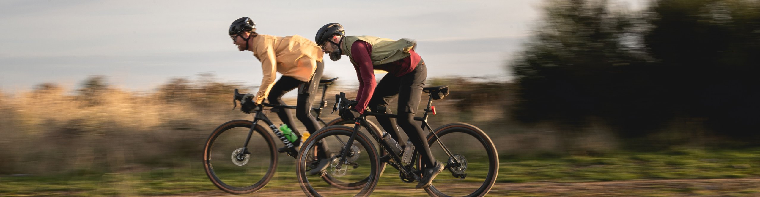 Zwei Gravelbiker rasen nebeneinander über einen Schottertrail, dynamisch in tiefer Rennposition, umgeben von herbstlicher Landschaft – Fokus auf Geschwindigkeit und Gravel-Performance
