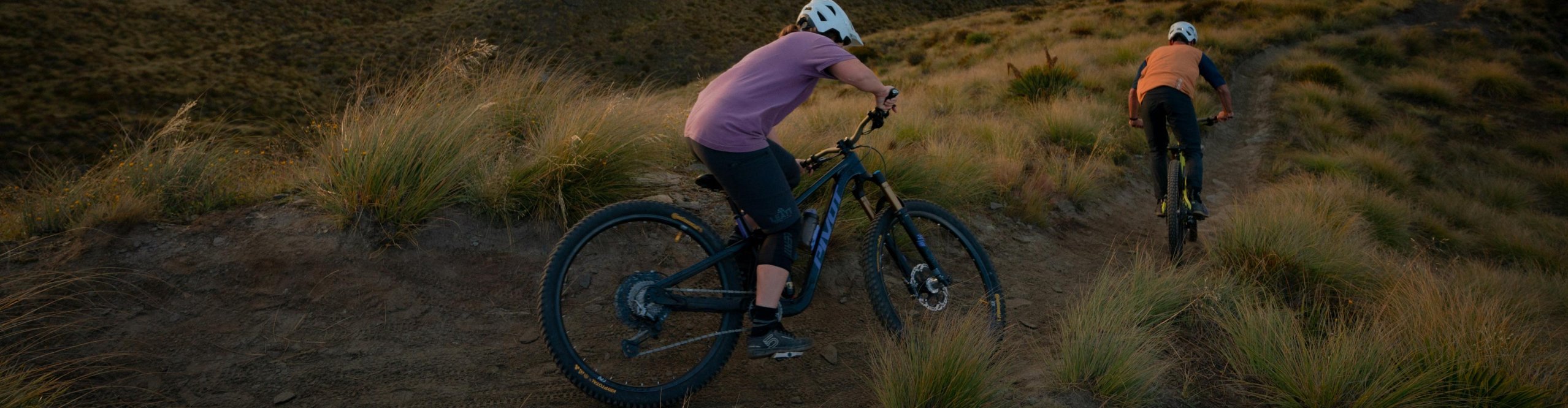 Two mountain bikers ride a trail at golden hour, featuring a Pivot Firebird enduro mountain bike in the foreground.